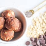 Almond Pulp Energy Balls in a white bowl surrounded by chia seeds, oats and chocolate chips.
