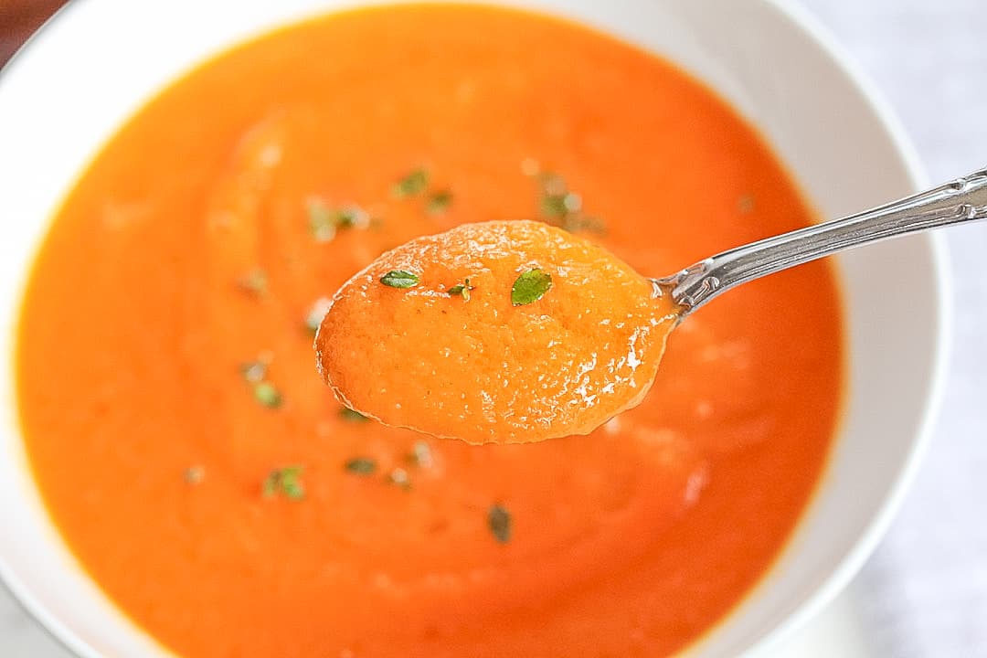 White bowl of tomato orange soup with a spoon scooping a bite.