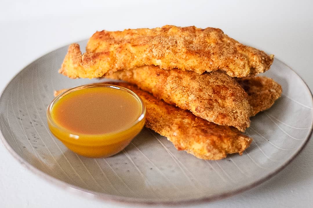 Pile of paleo chicken tenders on a plate with honey mustard dipping sauce in a small glass bowl.