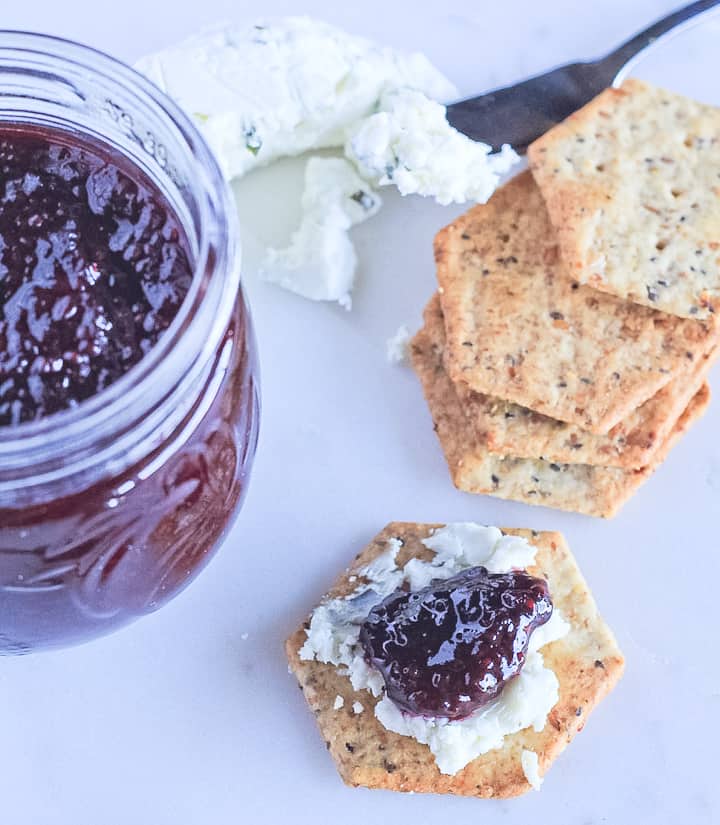A jar of blackberry chia seed jam, goat's cheese on a knife and a cracker topped with goats cheese and chia seed jam.