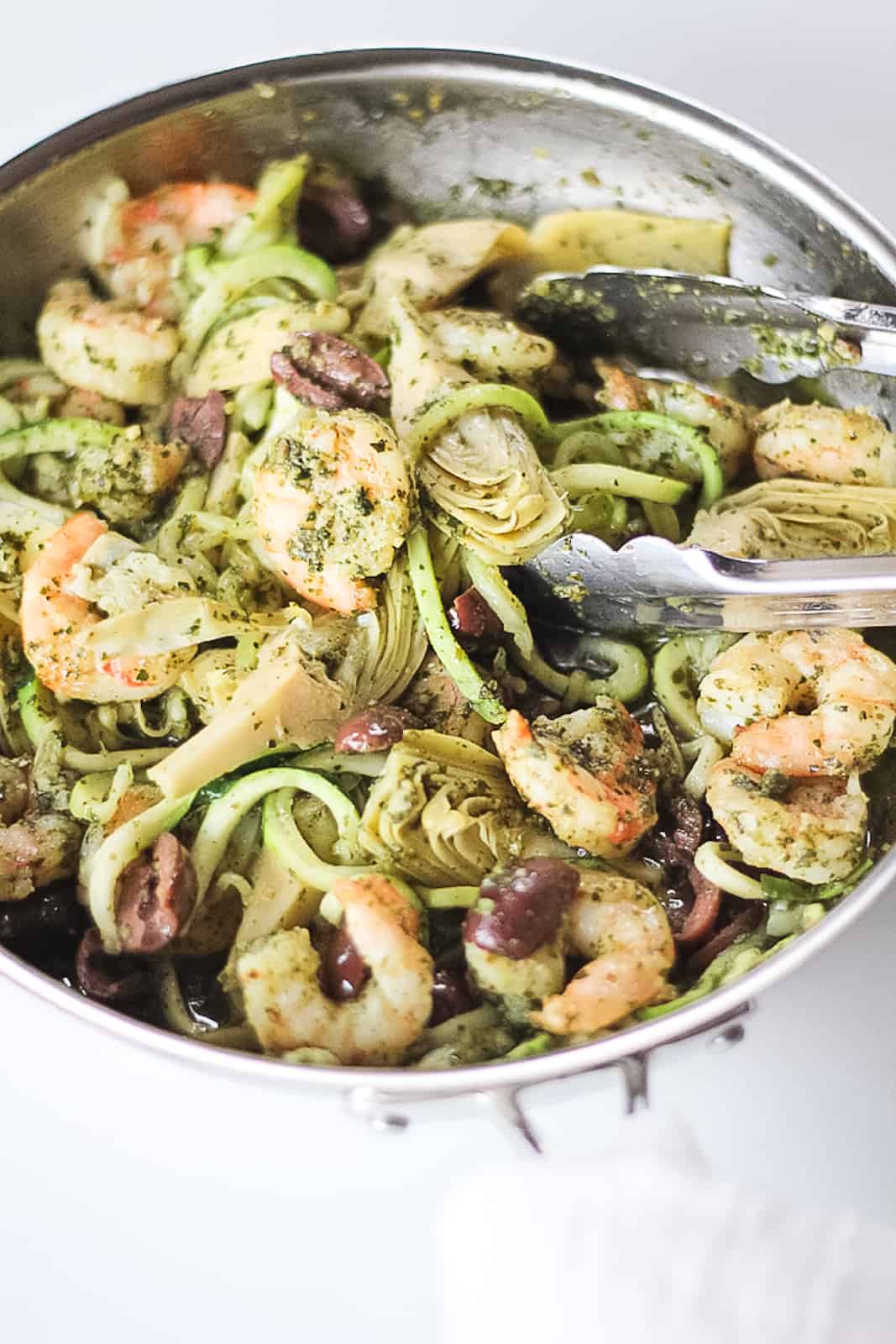Stainless steel pan full of pesto zucchini noodles, shrimp, artichoke hearts and olives for Pesto Zucchini Noodles with Shrimp recipe.