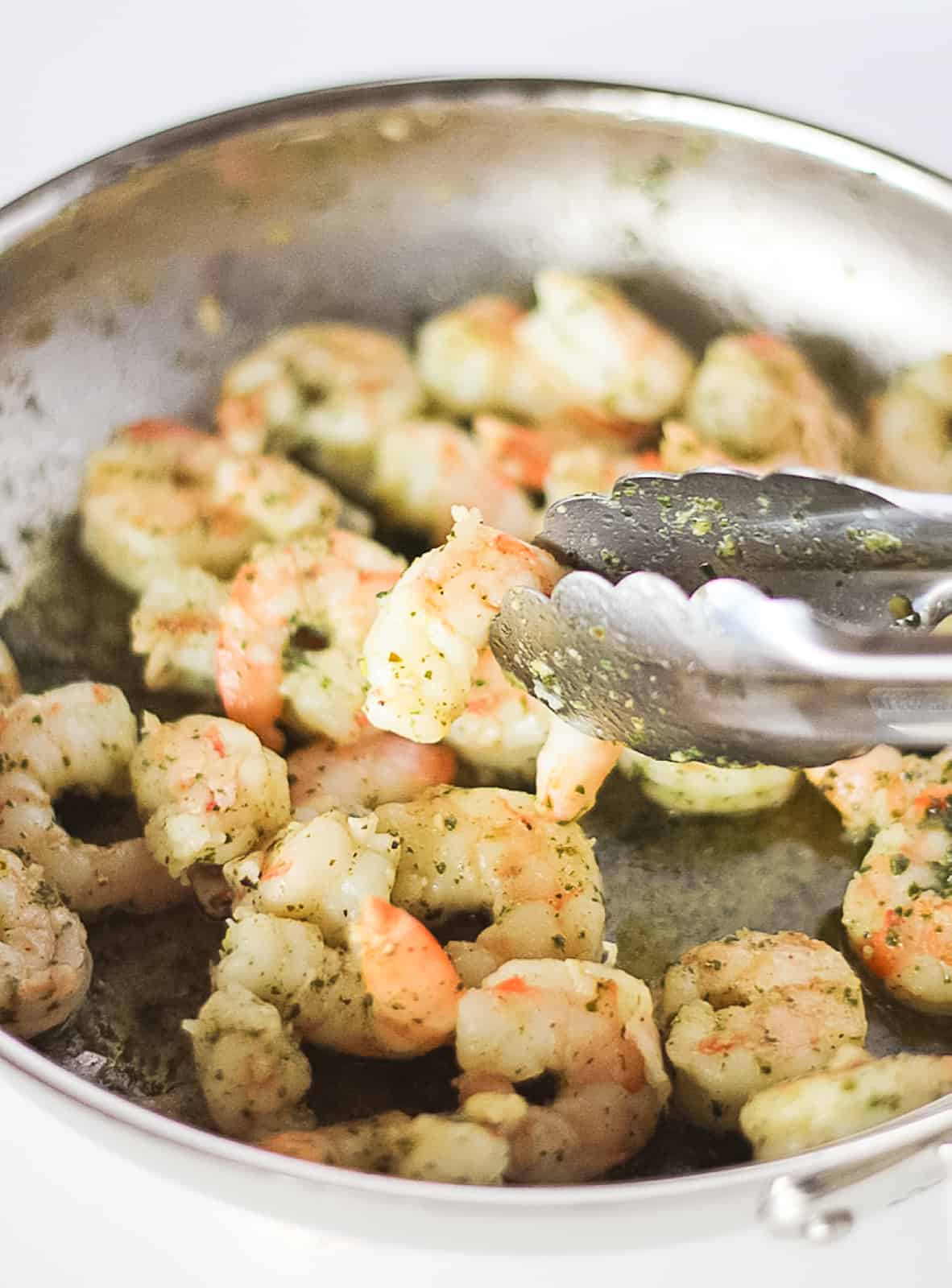 Sauted pesto shrimp in a stainless steel pan with tongs.