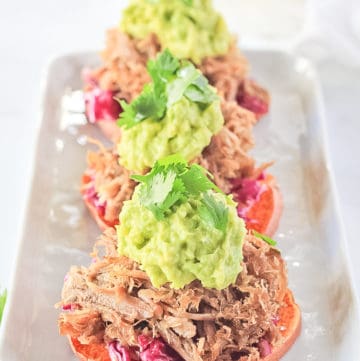 Carnitas Sweet Potato Sliders with lime cabbage, guacamole and cilantro on top a white plate.