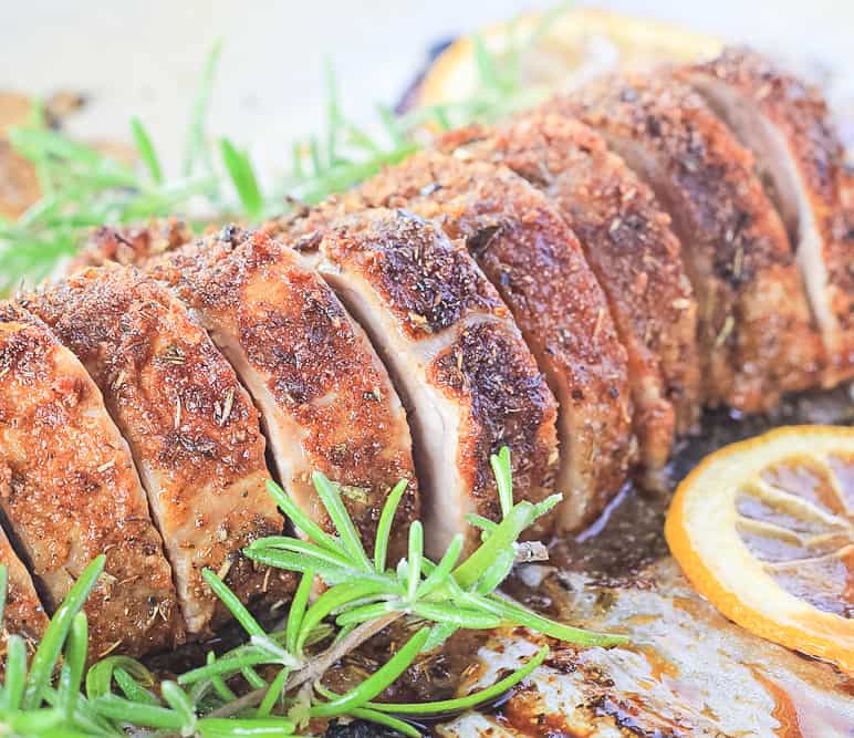 Slices of a pork tenderloin showing the spice rubbed crust on a sheet pan with lemon and rosemary sprigs.