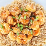 A white plate with orange shrimp over cauliflower rice garnished with sesame seeds and green onions.