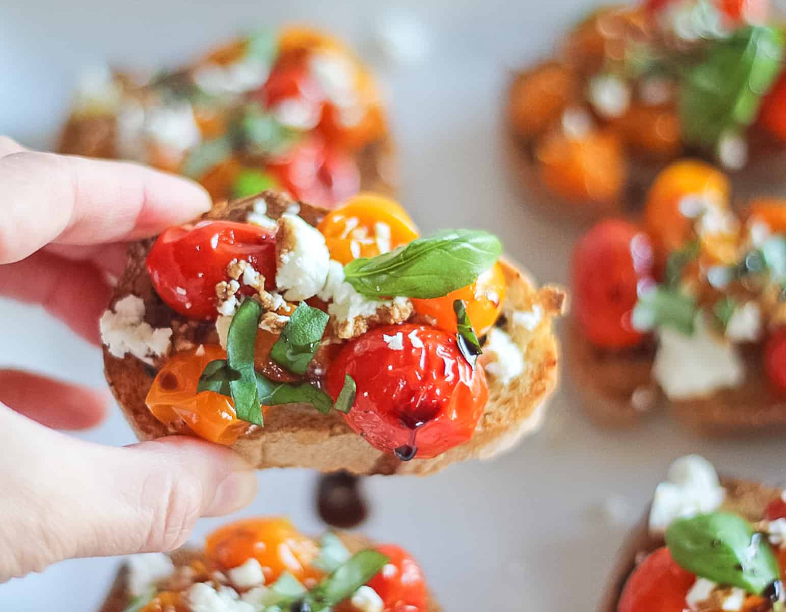 Hand picking up a cherry tomato, goat cheese, basil and balsamic appetizer.