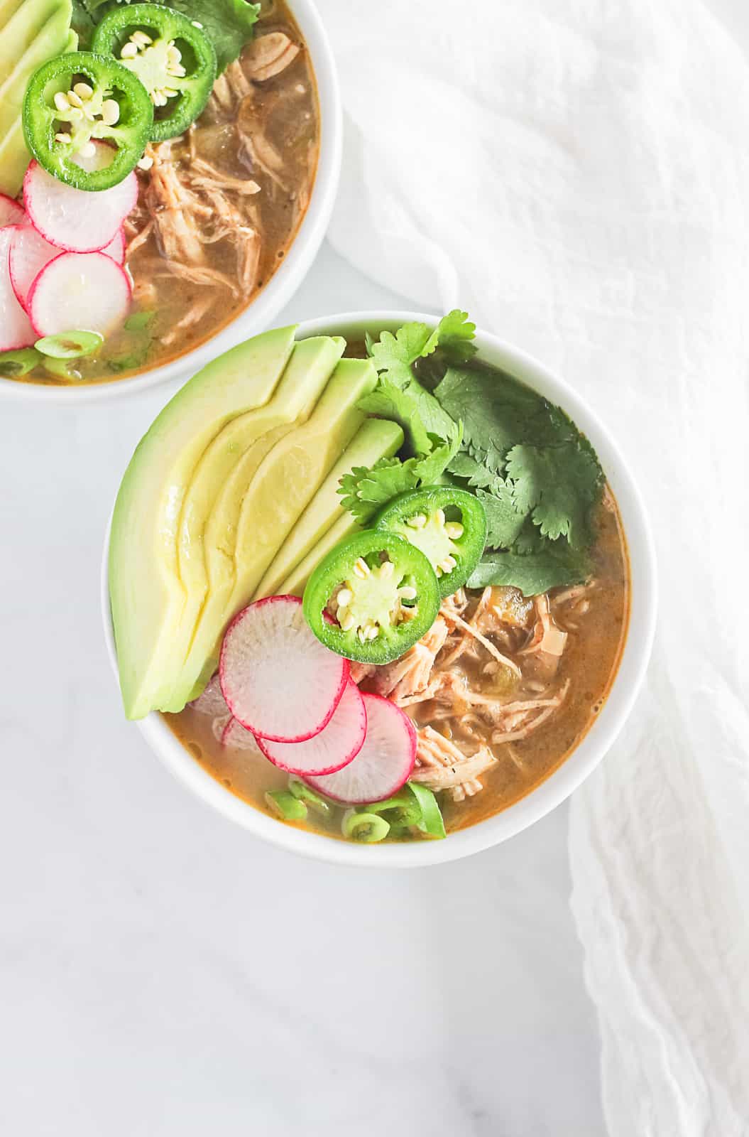 A white bowl of paleo white chicken chili topped with avocado, cilantro, jalapenos, radishes and green onion.