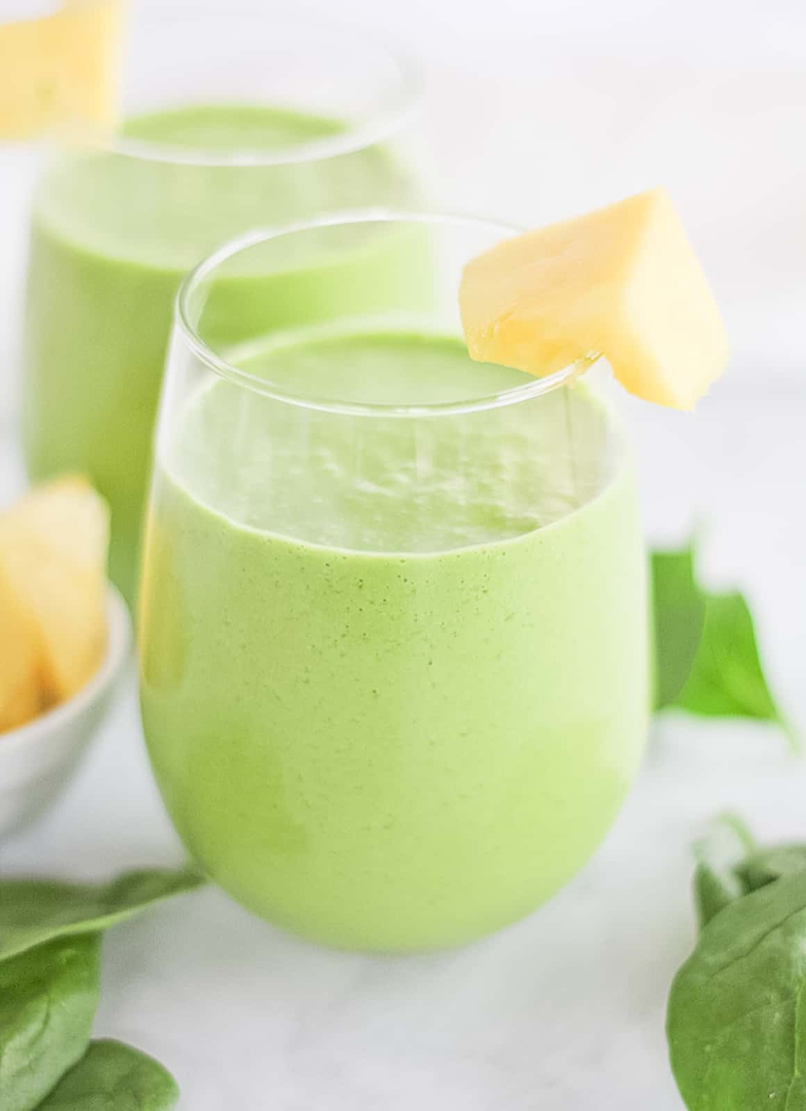 Close up of two glass cups filled with green smoothie surrounded by spinach leaves and chunks of pineapple.