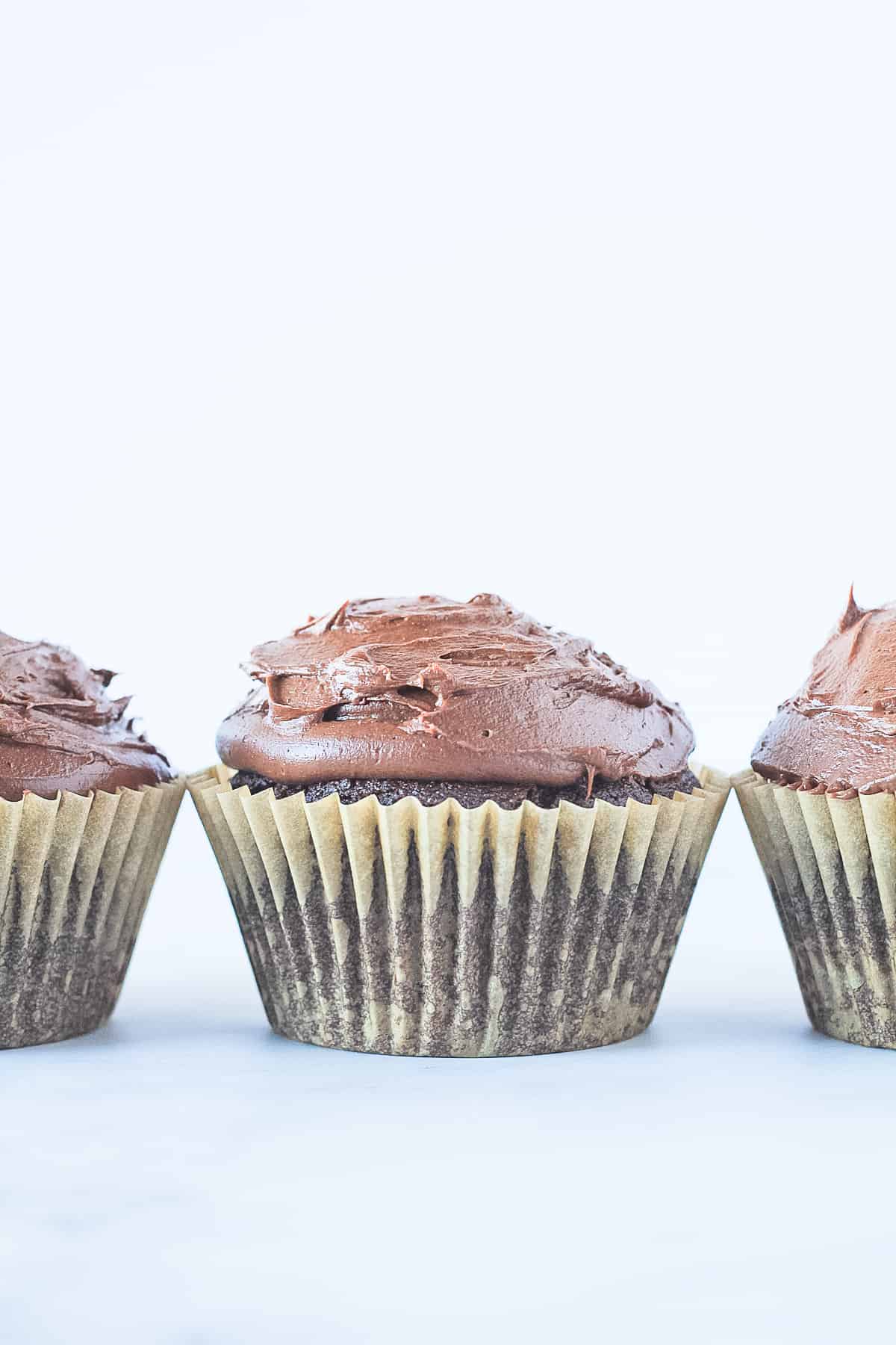 Three vegan chocolate cupcakes in a row topped with chocolate frosting.