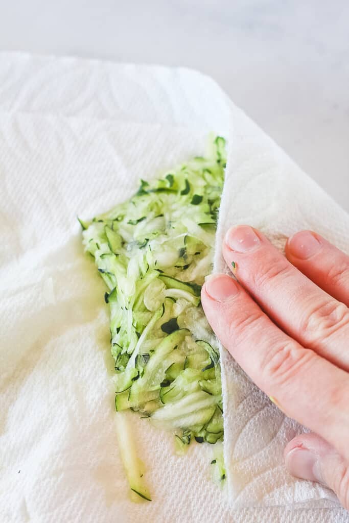 Hand pressing shredded zucchini with paper towel to remove moisture.