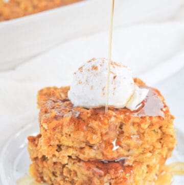 Two square of pumpkin baked oatmeal stacked on a white plate topped with a dollop of coconut cream, sprinkle of pumpkin pie spice and drizzle of maple syrup.