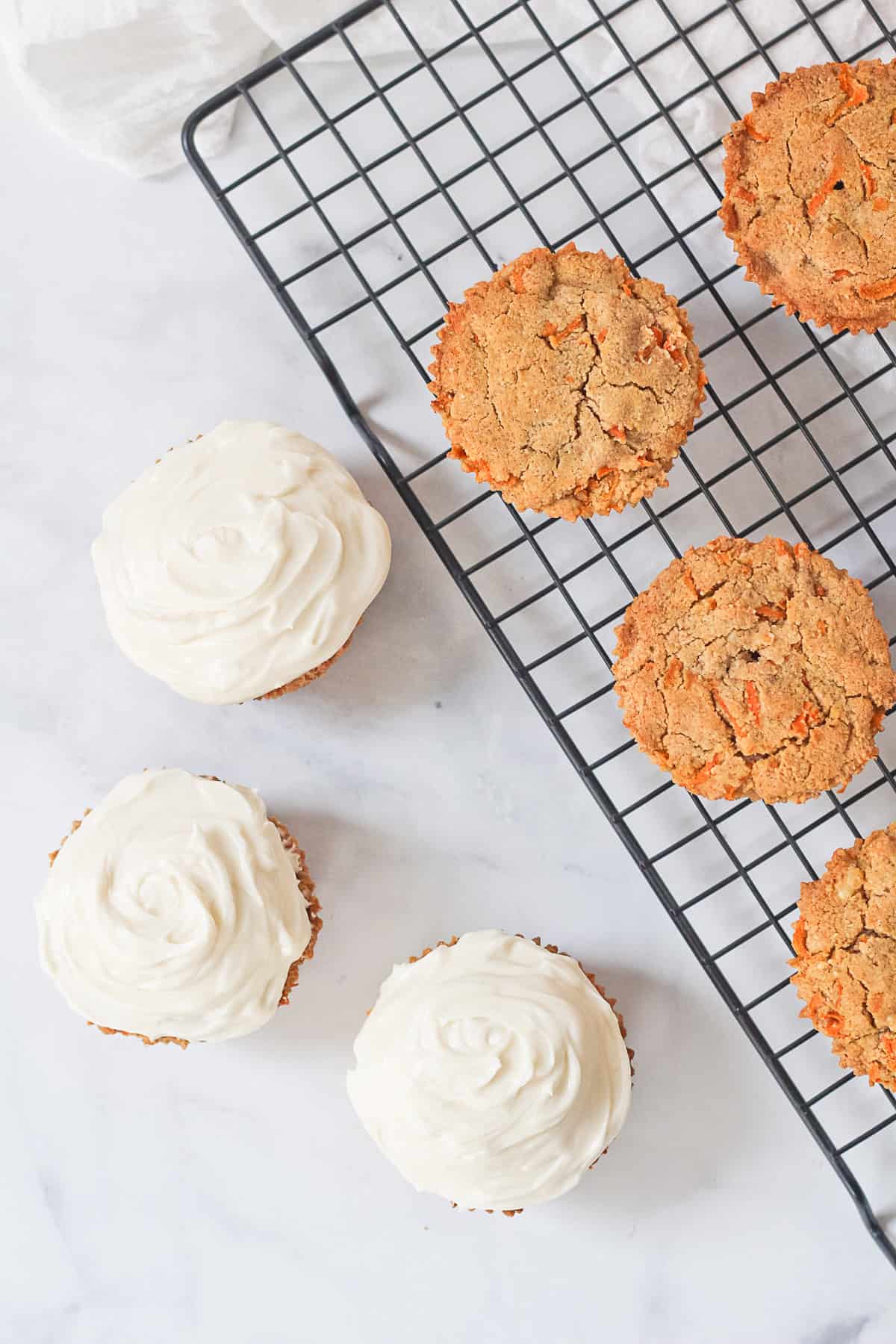 Three carrot cake muffins with vanilla frosting next to unfrosted muffins.